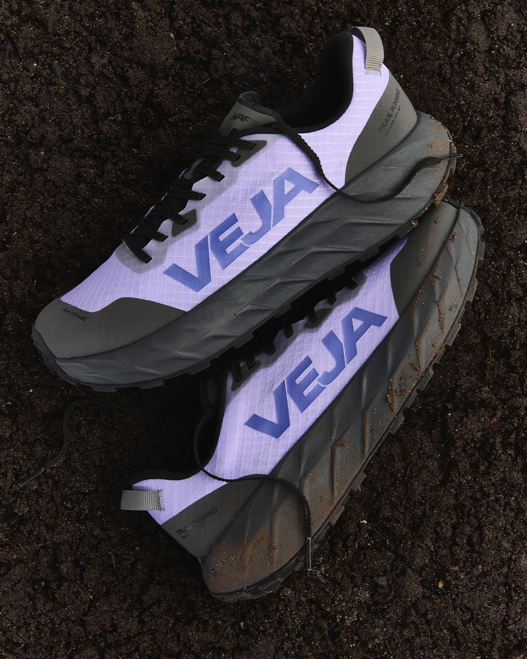 A pair of VEJA trail running shoes with a white and dark gray mesh upper, black laces, and a thick dark gray and black sole, placed on a bed of dark brown soil.