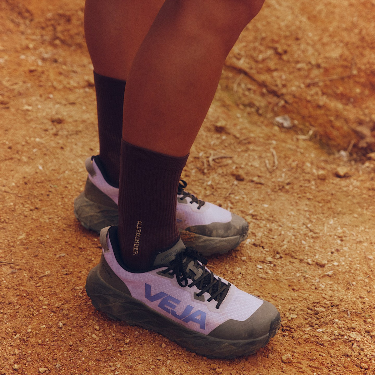 A close-up outdoor shot of a person's legs wearing black crew socks and trail running Veja Harpie Trail Aeroshell Lavande Acai sneakers on a sandy, dirt-covered path. The shoe has 'VEJA' printed on the side in blue.